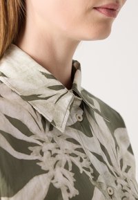 Chemise col montant dans des tons verts avec un motif floral. Présente une fermeture à boutons, un tissu texturé et un design décontracté.