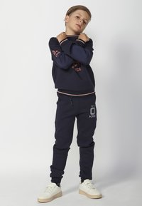 Sudadera navy con puños acanalados en contraste y detalle de logotipo, combinada con joggers a juego que presentan una impresión de logotipo. Las zapatillas son de color crema.