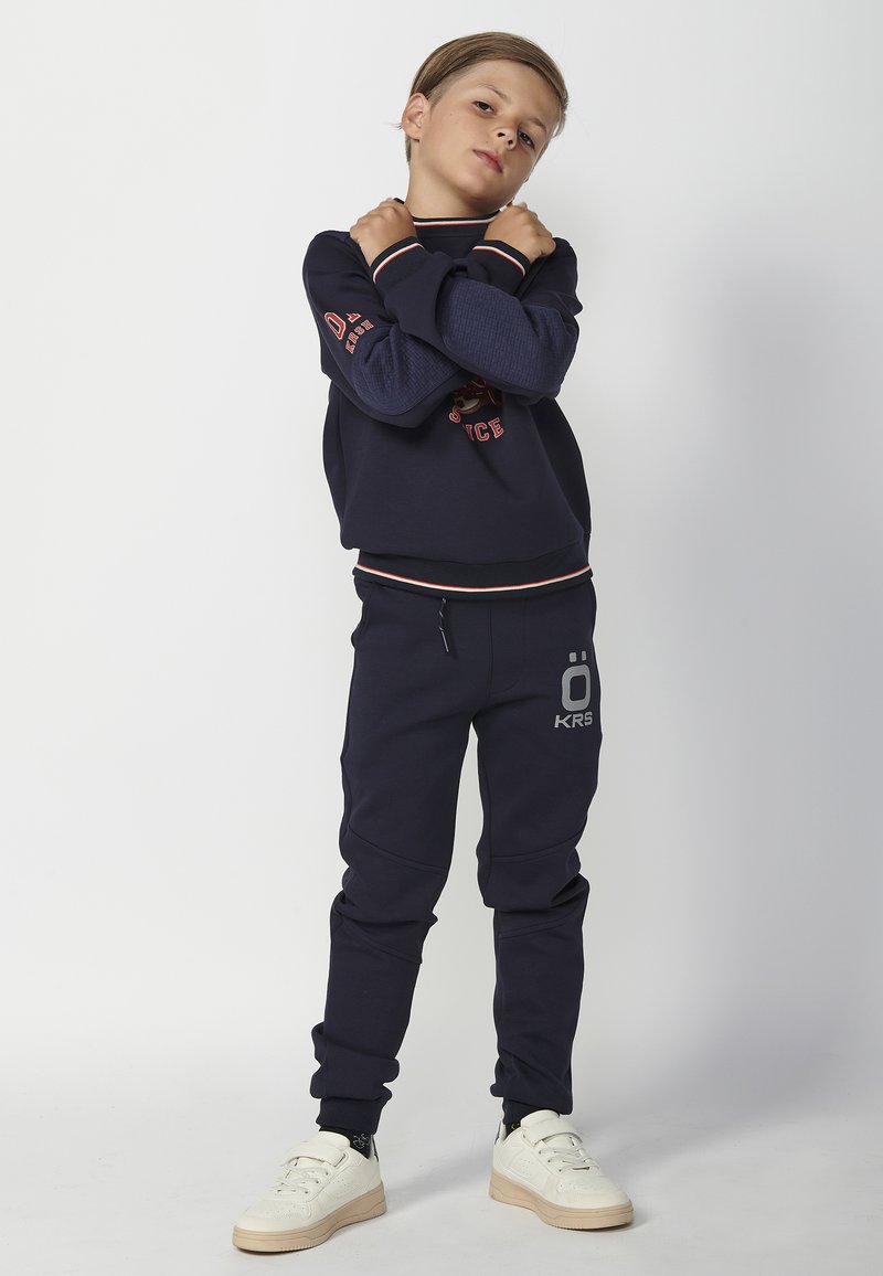 Sudadera navy con puños acanalados en contraste y detalle de logotipo, combinada con joggers a juego que presentan una impresión de logotipo. Las zapatillas son de color crema.
