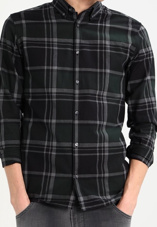 Homme portant une chemise à manches longues à carreaux noirs et gris, boutonnée, avec les mains dans les poches.