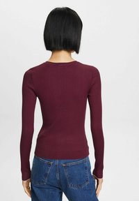 Syvä viininpunainen, uritettu pitkähihainen paita, vartalonmyötäinen malli pyöreällä kaula-aukolla ja hillityllä tekstuurilla, yhdistetty korkeavyötäröisiin farkkuihin.