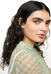 Top trasparente verde in rete con motivo a quadre, scollo alto e maniche lunghe. Modella con capelli ricci e orecchini a cerchio.