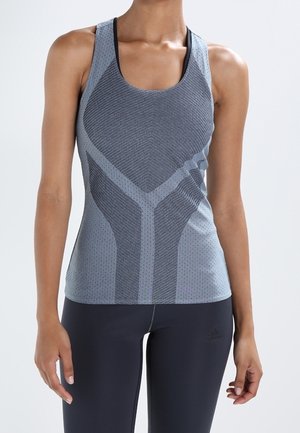 Femme portant un débardeur de sport bleu-gris ajusté avec une texture à motifs et un legging noir, debout devant un fond clair uni.