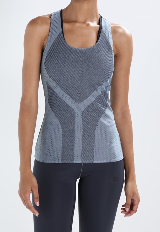 Femme portant un débardeur de sport bleu-gris ajusté avec une texture à motifs et un legging noir, debout devant un fond clair uni.