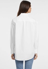 Camicia bianca a maniche lunghe con collo, polsini a bottoni e orlo curvo. Realizzata in tessuto liscio, mostrata dal retro. Design minimalista.