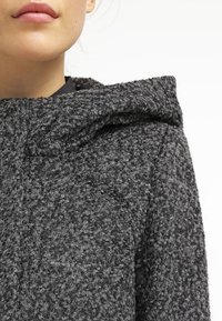 Felpa grigia con texture, dotata di un ampio cappuccio, caratterizzata da un tessuto ruvido e peloso. In un primo piano si possono vedere i dettagli della superficie e gli accenti delle cuciture.