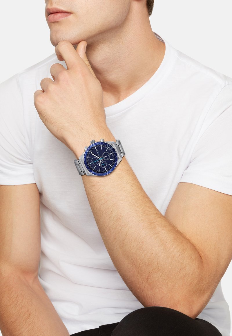 Homme portant un t-shirt blanc montrant une montre-bracelet en métal argenté avec un cadran bleu et des fonctionnalités de chronographe au poignet gauche.
