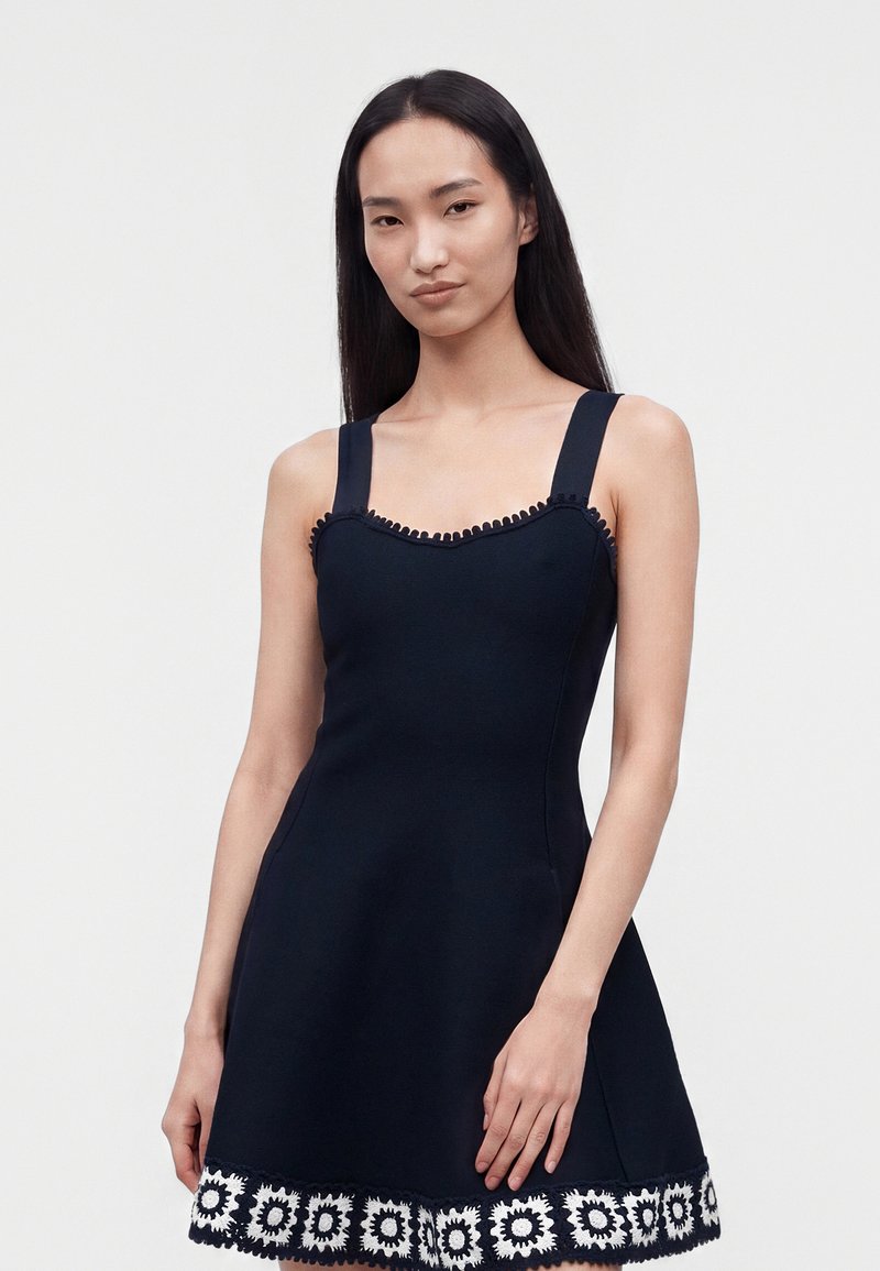 Jeune femme aux longs cheveux noirs portant une robe noire sans manches à larges bretelles avec une bordure florale blanche à l'ourlet, sur un fond uni.