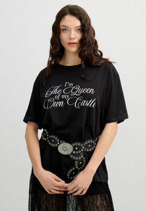 Joven con cabello rizado, vistiendo una camiseta negra que dice "Soy la reina de mi propio castillo" y un cinturón negro con ojales plateados.