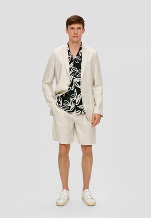 Lichtbeige linnen blazer, bijpassende shorts en zwart T-shirt met korte mouwen met een witte bladerenpatroon. Witte sneakers maken de look compleet.