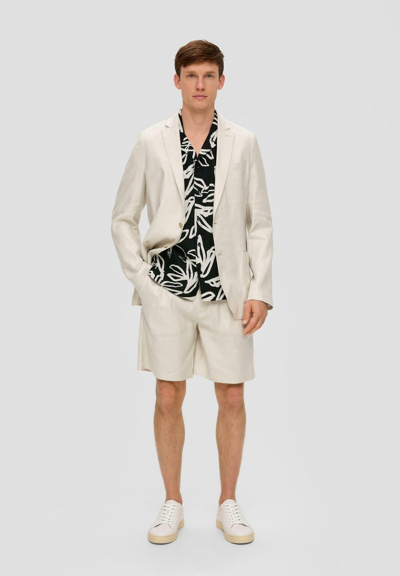 Lichtbeige linnen blazer, bijpassende shorts en zwart T-shirt met korte mouwen met een witte bladerenpatroon. Witte sneakers maken de look compleet.