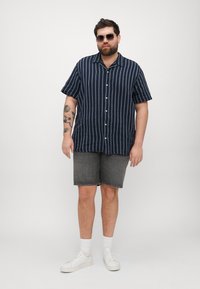 Camisa de rayas marinas de manga corta con botones, combinada con pantalones cortos de mezclilla grises y zapatillas blancas, de pie contra un fondo liso.