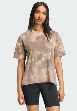 Woman with long dark hair wearing a brown tie-dye T-shirt and black fitted shorts standing against a light background.