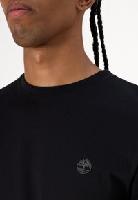 Sort bomulds t-shirt med rund hals, der har et lille gråt broderet trælogo på den øverste venstre side. Glat tekstur, tætsiddende design.