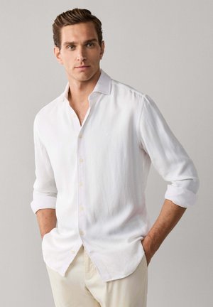 Hombre con el pelo marrón peinado, lleva una camisa blanca de manga larga abotonada y pantalones beige claros, manos en los bolsillos, mirando ligeramente hacia la derecha.