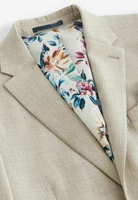 Blazer beige texturé avec doublure à motifs floraux dans des couleurs pastel. Présente un col cranté et une poche poitrine simple.