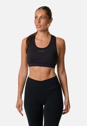 Femme portant un soutien-gorge de sport et un legging Swedemount noirs, debout, les mains détendues le long du corps, regardant légèrement vers sa gauche.