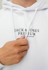 Weißer Hoodie mit verstellbaren Kordeln, ausgestattet mit einem schwarzen, bedruckten Logo "JACK & JONES PREMIUM" auf der Vorderseite und strukturiertem Stoff.