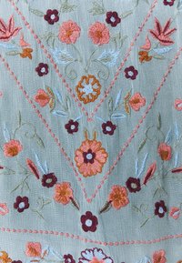 Broderat tyg med blommönster i rosa, orange och blå trådar på en ljusblå bakgrund, med sömnadsdetaljer i rosa.