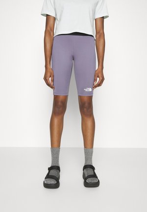 Persoon die paarse The North Face fietsbroeken, grijze sokken, zwarte sandalen en een wit cropped t-shirt draagt, staand tegen een witte achtergrond.