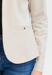 Ljusbeige blazer med en slät yta, som har en enkel sidoficka och en metallknapp. Matchad med mörkblå jeans.