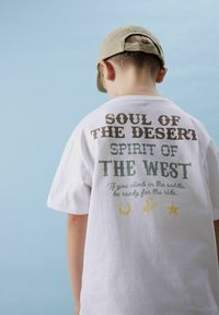 Valge puuvillane T-särk, mille seljal on erinevates värvustes tekst: "KÕRBE HING," "LÄÄNE VAIM," ning graafikad hobuseraua, tähe ja cowboy'ga.