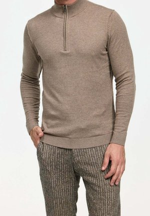 Pull zippé beige avec un col haut et des manches longues, fabriqué en tissu doux. Assorti avec un pantalon marron à motifs avec des rayures verticales.