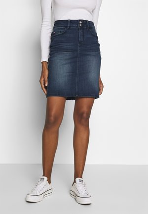 Femme portant une mini-jupe en jean bleu foncé, un haut blanc à manches longues et des baskets blanches, debout devant un fond blanc uni.