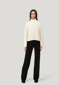 Cremefarbener, gerippter Strickpullover mit hohem Kragen und cropped Saum, kombiniert mit schwarzen, weit geschnittenen Hosen. Weiche Textur und entspannter Schnitt.