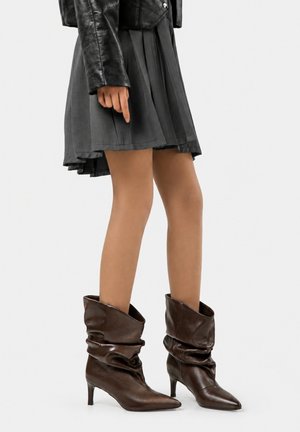 Frau trägt einen plissierten grauen Rock, schwarze Lederjacke und lässige dunkelbraune spitze Stiefel mit Absatz vor einfarbigem Hintergrund.