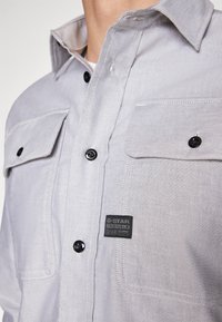 Harmaa nappipaita, jossa kaksi rintataskua, mustat napit ja merkkilätkä. Kangas on sileää ja siinä on hienovaraisia ompeluvaria.