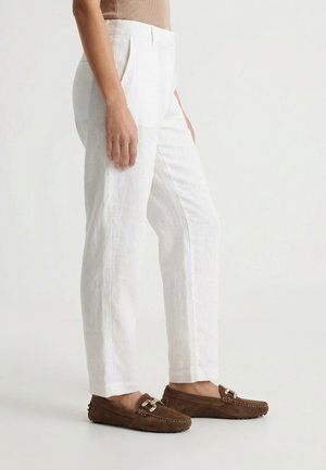 Persoon draagt witte linnen broek en bruine suède loafers met gouden gespsdetails, staand op een lichtgrijze vloer.