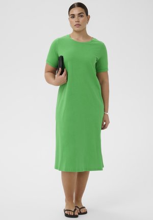 Kvinde i en grøn knælængde kjole med korte ærmer, der holder en sort clutch med den ene hånd, iført sorte flip-flops og et armbåndsur.