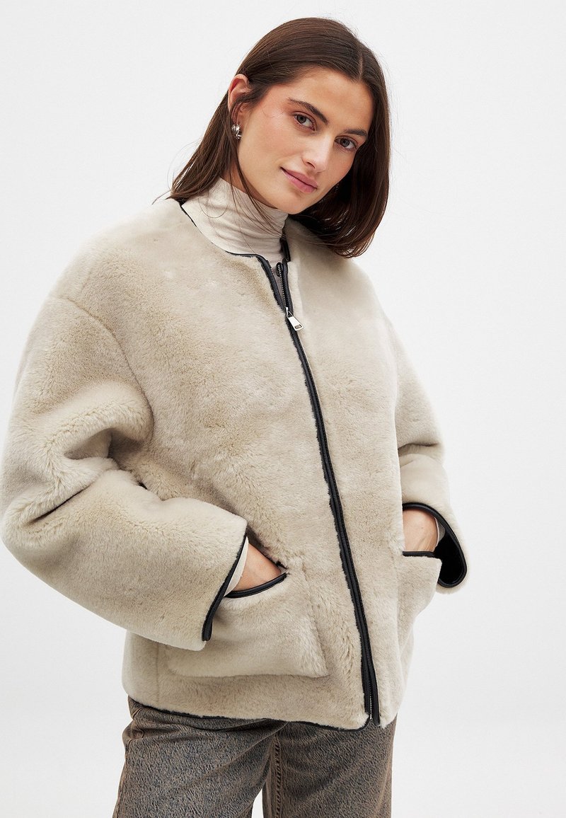 Bēšas mākslīgās kažokādas jaka ar melnu rāvējslēdzēju, apaļu izgriezumu un divām sānu kabatām. Mīksta tekstūra un brīvas piegriezuma dizains.