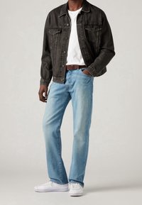 Giacca in denim nera con taglio classico, t-shirt bianca, jeans azzurri chiari e sneakers bianche. Caratteristiche includono tasche sul petto e una cintura marrone.