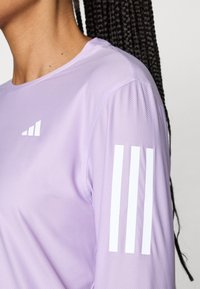 Fialové sportovní tričko s texturovanou látkou, kulatým výstřihem a bílým trojpruhovým logem na levém rukávu.