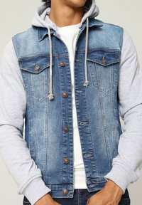 Veste en denim avec des manches de sweat à capuche grises, deux poches sur la poitrine, des boutons marron et une texture tissée. Présente des détails usés sur le denim.