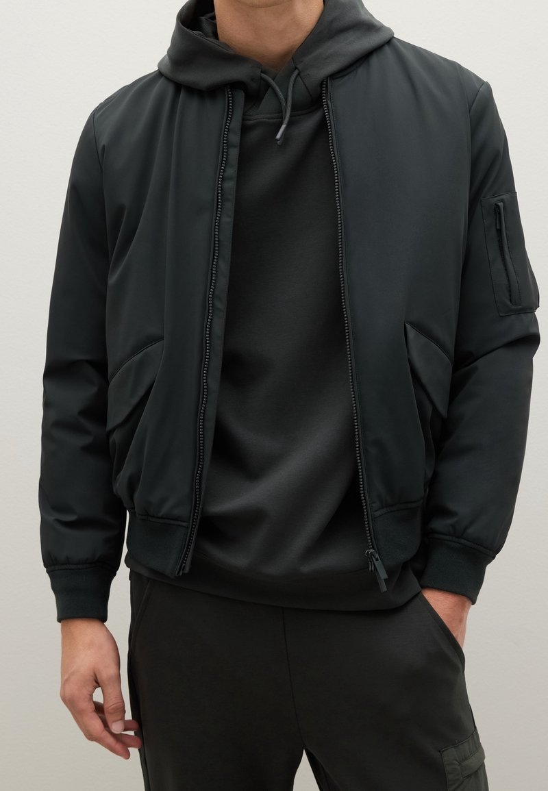 Homme portant une veste bomber noire à fermeture éclair sur un sweat à capuche noir et un pantalon noir, debout avec une main dans la poche devant un fond uni.