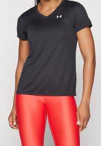 Svart kortärmad träningströja med v-ringning, slät textur och en vit logo på vänster bröst. Kombinerad med glänsande röda tights.