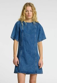 Blå denimkjole med en løs passform, korte ermer og en rett silhuett. Avsluttet med synlige sømmer og rå nederkant.