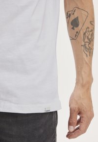 Camiseta de algodón blanca con corte recto, con una pequeña etiqueta de tela en el dobladillo. Mano que muestra tatuajes y un diseño de carta de juego.