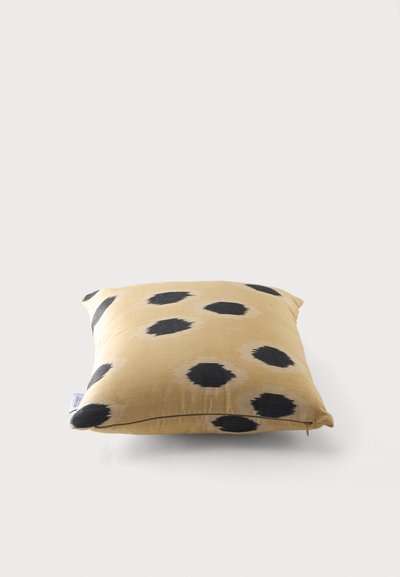 Coussin carré beige avec des taches noires irrégulières dispersées sur son tissu, reposant sur une surface blanche unie.