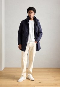 Giacca puffer blu navy con colletto alto, chiusura a bottoni sul davanti e tasche laterali, abbinata a pantaloni beige e sneakers bianche su un pavimento di legno.
