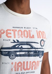 Valkoinen puuvillapaita, jossa on kuvio siniraitaisesta autosta ja punaisista teksteistä "PETROL IND" ja "HAWAII", retrotyylillä.