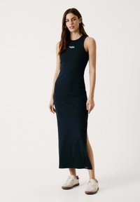 Navyblaues, ärmelloses Maxikleid aus weichem Stoff mit seitlichen Schlitzen, versehen mit einem kleinen weißen Logo auf der Brust und kombiniert mit weißen Sneakers.