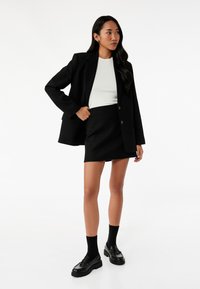 Blazer negro ajustado sobre una blusa blanca, combinado con una falda mini texturizada negra. La modelo lleva calcetas negras y mocasines negros gruesos.