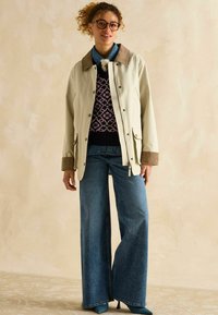 Jachetă waterproof bej deschis, cu guler maro, asortată cu un pulover bleumarin cu model și blugi evazați bleu. Pantofi cu toc înalt teal.