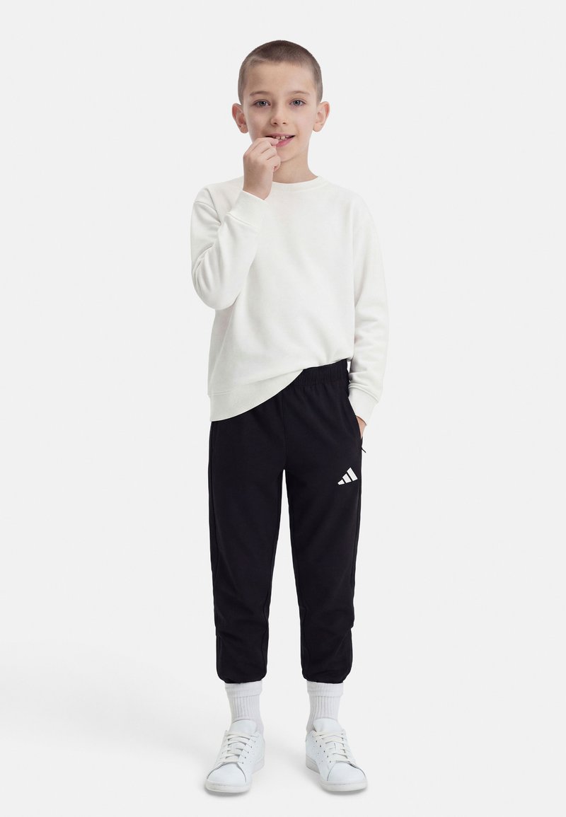 Αγόρι με λευκή φούτερ, μαύρο παντελόνι φόρμας Adidas, λευκές κάλτσες και αθλητικά παπούτσια, στέκεται με το ένα χέρι στην τσέπη και το άλλο κοντά στο στόμα.