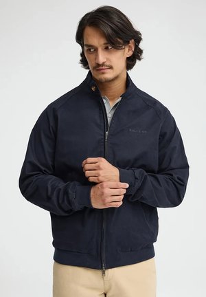Uomo che sistema il polsino di una giacca blu navy con zip sopra una camicia grigio chiaro, indossando pantaloni beige, in piedi davanti a uno sfondo semplice.