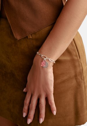 Arm mit einem klobigen silbernen Kettenarmband, verziert mit einem rosa Gummibärchen und runden silbernen Anhängern, vor einem braunen Wildlederrock und Oberteil.